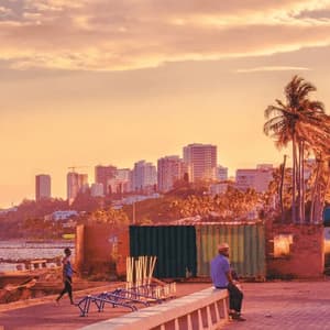 A man sits on a concrete ledge on a waterfront, looking out at a modern city skyline with palm trees during a warm sunset.