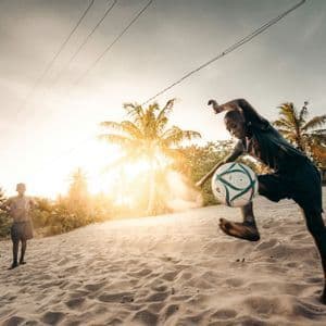 A young boy kicks a football in the air on a sandy beach at sunset, with another boy and palm trees in the background.