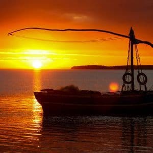 A silhouetted boat floats on calm water during a vibrant orange sunset over the sea.