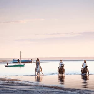A WeRoad group trip with three women riding horses through the shallow water along a beach during a calm sunset.