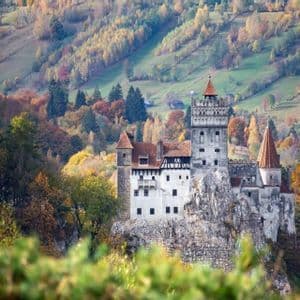 A medieval castle with red-tiled towers stands on a rocky cliff surrounded by hills covered in vibrant autumn trees.