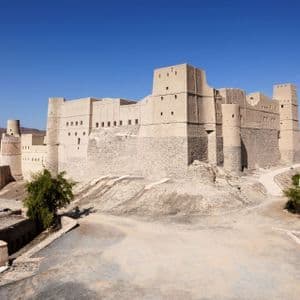 Una grande fortezza color sabbia con torri sorge su una collina che domina una città e montagne sotto un limpido cielo azzurro.