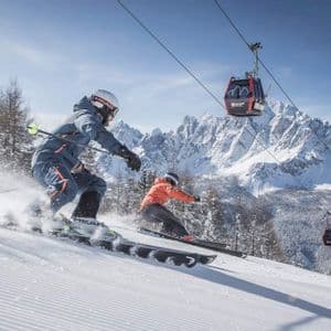 Two people from a WeRoad group trip skiing down a snowy slope, with a cable car overhead and mountains in the background.