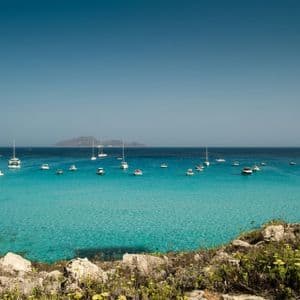 Una vista da una costa rocciosa di numerose barche ancorate in un mare calmo e turchese sotto un cielo azzurro e limpido, con un'isola in lontananza.