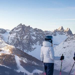 From behind, two skiers from a WeRoad group trip pause to look at a sunlit, snow-covered mountain range.