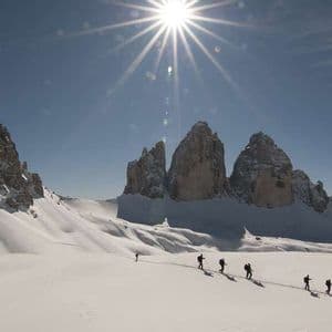 A WeRoad group trip hikes in a single file line across a vast snowy mountain landscape under a bright, sunny sky.