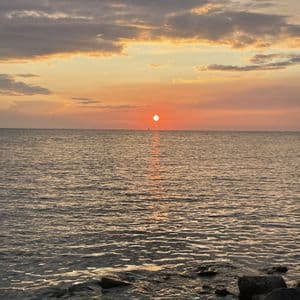 Il sole tramonta all'orizzonte sull'acqua, tingendo le nuvole di un bagliore arancione e riflettendosi sulla superficie increspata vicino a una riva rocciosa.