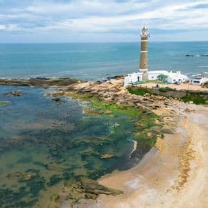 Veduta aerea di un alto faro in pietra che si erge su una costa rocciosa accanto a una caletta sabbiosa con acqua limpida.