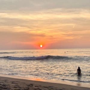Une personne se tient dans l'eau peu profonde de l'océan au coucher du soleil, tandis que plusieurs surfeurs attendent les vagues au loin.