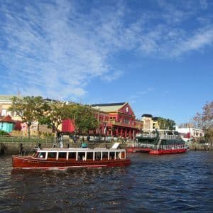 Una barca turistica in legno naviga su un fiume passando davanti a un lungomare con edifici colorati e alberi sotto un cielo azzurro.