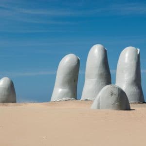 Grande scultura bianca di dita che affiorano dalla sabbia della spiaggia sotto un cielo azzurro.