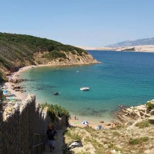 A high-angle view of a sunny coastal cove with turquoise water, a sandy beach with people, and boats floating near the shore.