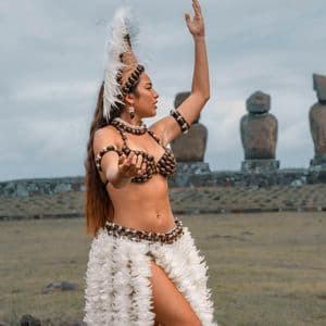 Una donna in gonna bianca piumata e copricapo tradizionale danza in un prato erboso con grandi statue di pietra sullo sfondo.