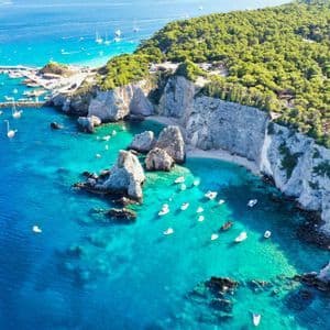 Vista aerea di una costa rocciosa con ripide scogliere bianche e una foresta verde, dove numerose piccole imbarcazioni galleggiano sul mare turchese.