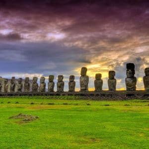 Una fila di grandi statue Moai in pietra, erette su una piattaforma in un campo erboso, sotto un cielo drammatico e nuvoloso al tramonto.