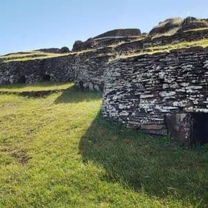 Antiche case in pietra a secco con piccoli ingressi si integrano in un pendio erboso terrazzato, sotto un cielo azzurro.