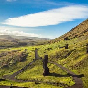 Grandi statue Moai sono sparse su un pendio erboso con un sentiero tortuoso sotto un cielo parzialmente nuvoloso.