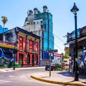 Un angolo di strada cittadino soleggiato con graffiti colorati sui palazzi, accanto a un moderno grattacielo di vetro.