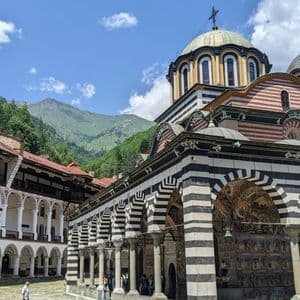 Un monastère orné avec des arches rayées et de grands dômes se dresse dans une cour avec des montagnes vertes en arrière-plan sous un ciel partiellement nuageux.