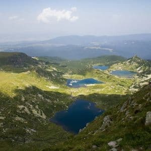 Vue plongeante d'un groupe WeRoad en randonnée sur un sentier rocheux, surplombant une vallée verdoyante parsemée de plusieurs lacs de montagne d'un bleu profond.