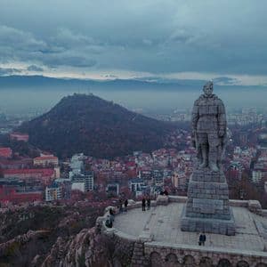 Un grand monument en pierre d'un soldat se dresse sur une colline surplombant une ville tentaculaire avec des montagnes lointaines sous un ciel nuageux.