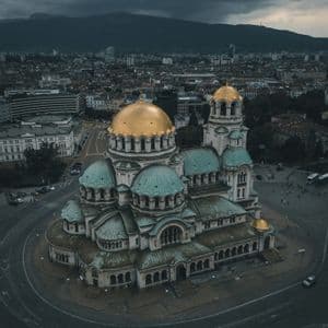 Une vue aérienne d'une grande cathédrale avec des dômes or et verts, entourée d'un paysage urbain, avec des montagnes sous un ciel nuageux.