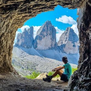 Eine Person sitzt am Eingang einer Höhle und blickt auf drei zerklüftete Berggipfel unter blauem Himmel mit Wolken.