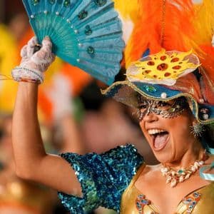 Eine lächelnde Frau in einem farbenfrohen Karnevalskostüm mit Federkopfschmuck und Maske hält einen blauen Fächer in die Höhe.