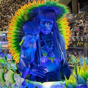 Ein großer, blauer Karnevalswagen mit einer indigenen Frau und einem Kind mit farbenprächtigem Federkopfschmuck, bei einem Umzug vor einer großen Menschenmenge.