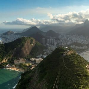 Ein Blick aus der Seilbahn auf eine Küstenstadt, eingebettet zwischen üppig grünen Bergen und dem Ozean unter einem teils bewölkten Himmel.