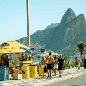Menschen in Sommerkleidung spazieren auf einer gemusterten Strandpromenade an einem gelben Kiosk vorbei, im Hintergrund erheben sich große grüne Berge unter einem klaren Himmel.
