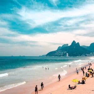 Ein weitläufiger Blick auf einen belebten Strand, an dem Menschen schwimmen und unter Sonnenschirmen entspannen, mit hohen Bergen, die sich hinter einer Küstenstadt erheben.