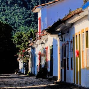 Eine sonnenbeschienene Kopfsteinpflasterstraße, gesäumt von weißen Gebäuden im Kolonialstil mit bunten Türen und Fenstern, mit einem üppig grünen Berghang im Hintergrund.