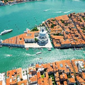 Vista aerea di una città con tetti di tegole arancioni e un grande edificio a cupola bianco circondato da acqua turchese e barche.