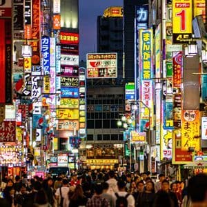 Una folla di persone cammina lungo una stretta strada cittadina al crepuscolo, illuminata dal bagliore di numerose e colorate insegne al neon giapponesi sugli edifici.