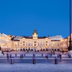 Una vasta piazza cittadina, circondata da edifici storici illuminati, sotto un cielo blu intenso al crepuscolo.