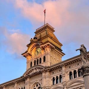 Uno scatto dal basso di un'ornata torre dell'orologio su un edificio storico, contro un cielo con nuvole rosa al tramonto.