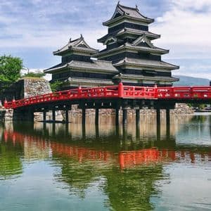 Un castello giapponese tradizionale con un ponte rosso brillante di fronte, entrambi riflessi nelle calme acque di un fossato sotto un cielo nuvoloso.