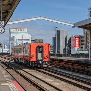 Un treno arancione sui binari su una banchina di stazione all'aperto con edifici cittadini visibili sullo sfondo sotto un cielo azzurro.