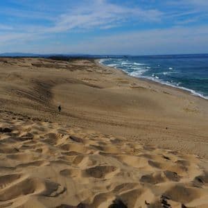 Vaste dune di sabbia che conducono a una costa oceanica blu, con alcune figure distanti che camminano sotto un cielo parzialmente nuvoloso.