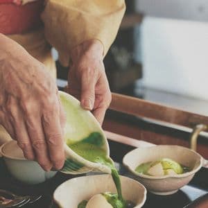 Una persona in kimono giallo versa salsa matcha verde da una ciotola di ceramica su palline di gelato alla vaniglia.