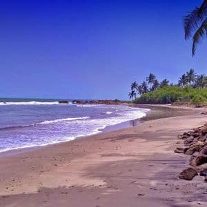 Le onde dell'oceano lambiscono una spiaggia sabbiosa costeggiata da rocce e palme sotto un cielo limpido e di un blu profondo.