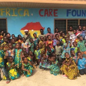 Un folto gruppo di bambini e alcuni adulti con abiti colorati posano per una foto davanti a un edificio blu con scritto 'Africa Care Foundation'.