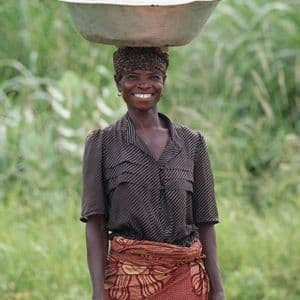 Una donna sorridente regge in equilibrio una grande ciotola di frutta sulla testa, all'aperto, davanti a dell'erba alta e verde.