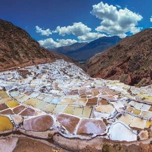Una vista panorámica de salinas en terrazas de varios colores anidadas en un valle de montaña bajo un cielo azul y nublado.