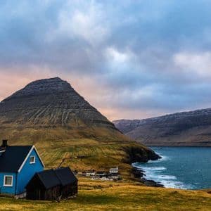 Una casa blu su una collina erbosa si affaccia su uno specchio d'acqua e una grande montagna sotto un cielo nuvoloso.