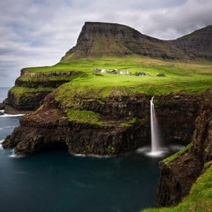 Una cascata si getta da una scogliera erbosa nell'oceano sottostante, con un piccolo villaggio costiero e montagne sotto un cielo nuvoloso.