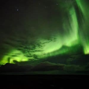 La vivace aurora boreale verde illumina il cielo notturno, serpeggiando tra nuvole e stelle su un paesaggio oscuro.