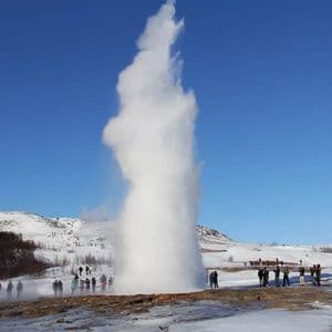 Un viaggio di gruppo WeRoad ammira un grande geyser che erutta in un paesaggio innevato sotto un cielo azzurro e limpido.