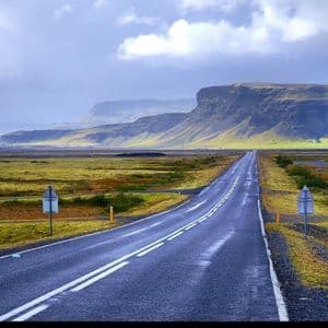 Una lunga strada dritta si estende attraverso un vasto paesaggio muschioso verso montagne distanti e dalla cima piatta sotto un cielo nuvoloso.